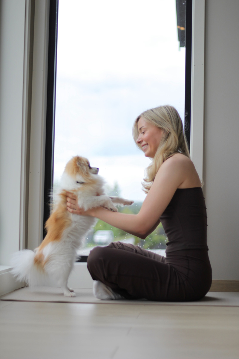 Malin Kristine Moen sitter ved vinduet og holder den lille hunden sin, som strekker seg opp mot henne, i et lyst og rolig rom.